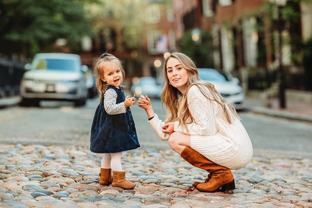 A young woman in a white dress and brown boots crouches on a cobblestone street, smiling while holding hands with a happy toddler wearing a blue dress and brown boots—an enchanting moment that captures the joy of preparing kids for photoshoot memories.