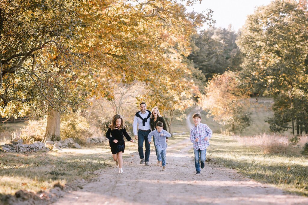 A family of five walks and runs along a sunlit dirt path in a park with autumn trees, smiling and enjoying a pleasant day outdoors after preparing their kids for the photoshoot.