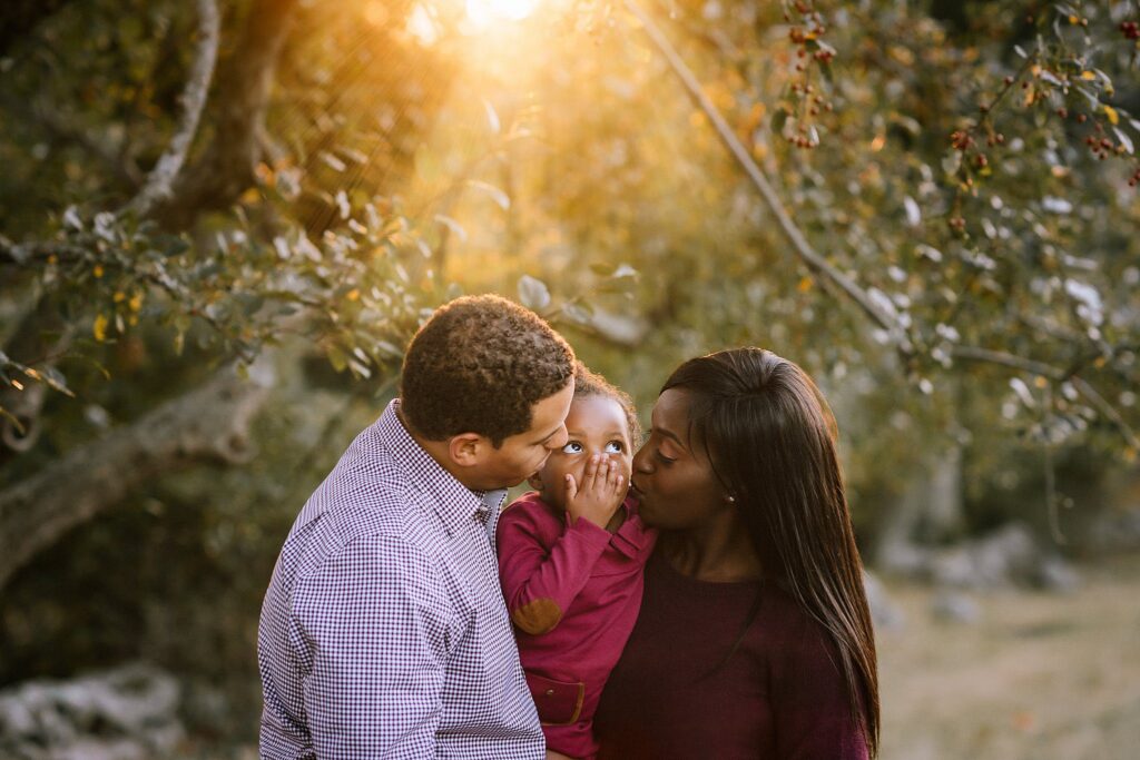 A family stands outdoors at sunset, clearly enjoying the moment. A man and woman smile at a small child between them. The child, wearing a maroon top, covers their mouth with their hands in a candid gesture. Sunlight streams through the trees in the background.