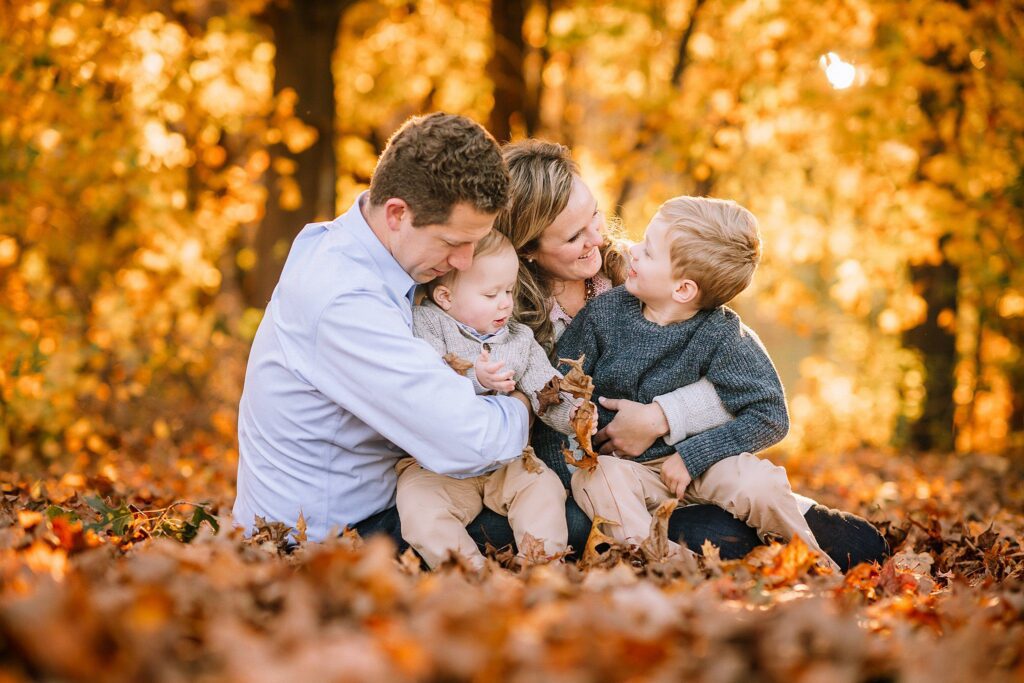 A family of four sits together in a pile of autumn leaves, smiling and laughing, surrounded by trees with golden foliage—thanks to preparing kids for photoshoot moments like this, their joy shines through naturally.