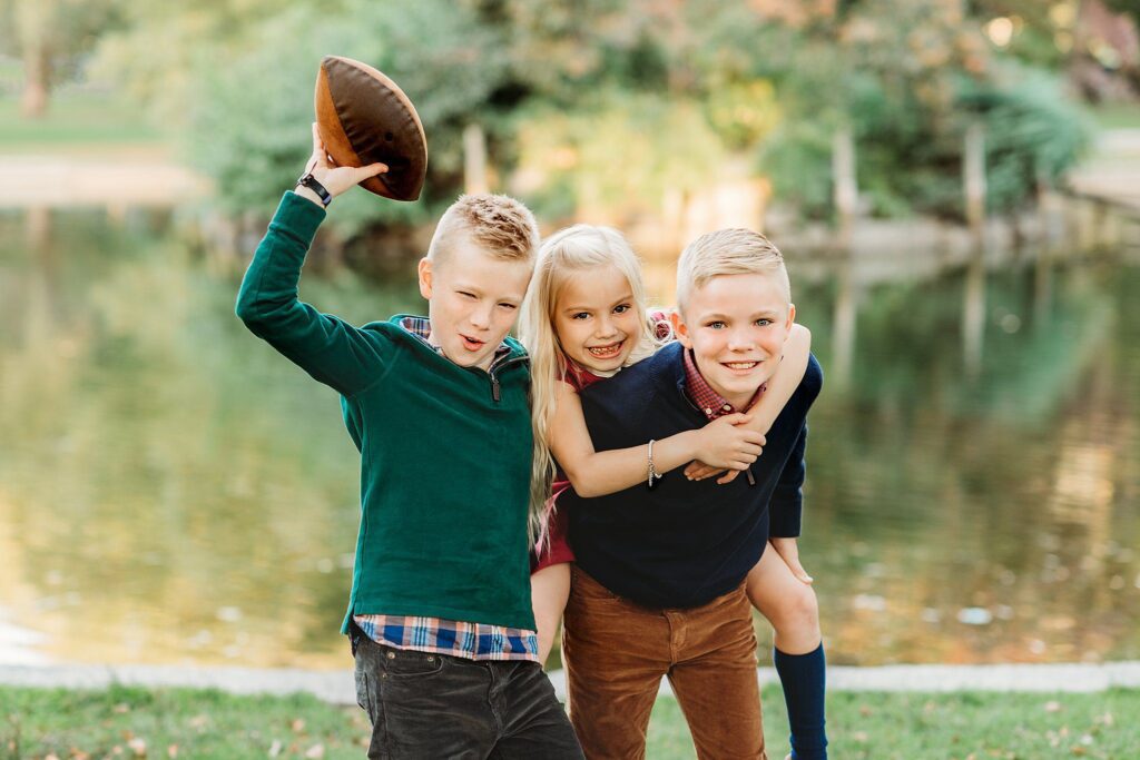 Three smiling children pose by a pond; one boy holds a football, a girl hugs both boys from behind, and the other boy bends forward. They have cheerful expressions and natural poses. They wear casual clothes and stand on grass with trees in the background.