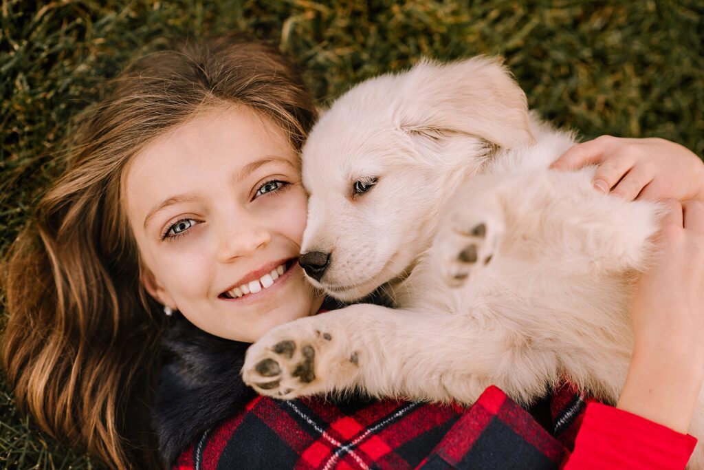 A smiling young girl lies on grass, cuddling a fluffy white puppy in her arms. Both appear happy and content, capturing the magic that comes from preparing kids for a photoshoot with their favorite pets.