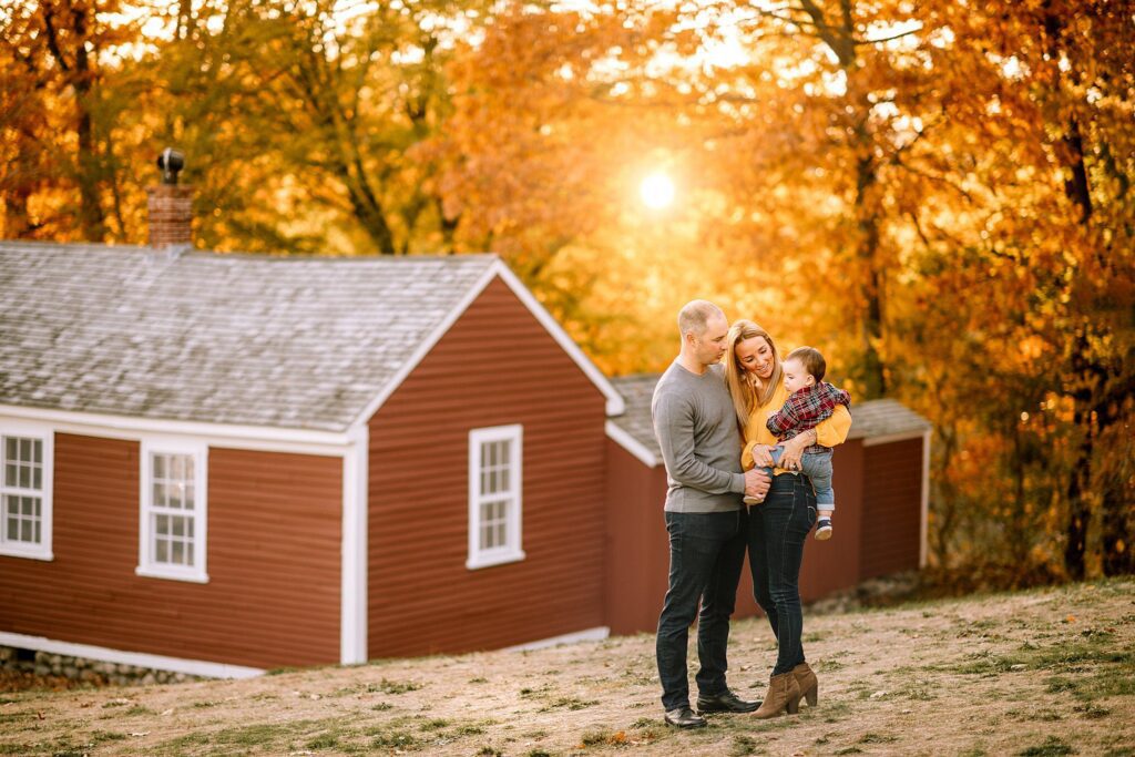 A couple stands outside holding a baby, with autumn trees and a red house in the background as the sun sets, casting a warm golden light.