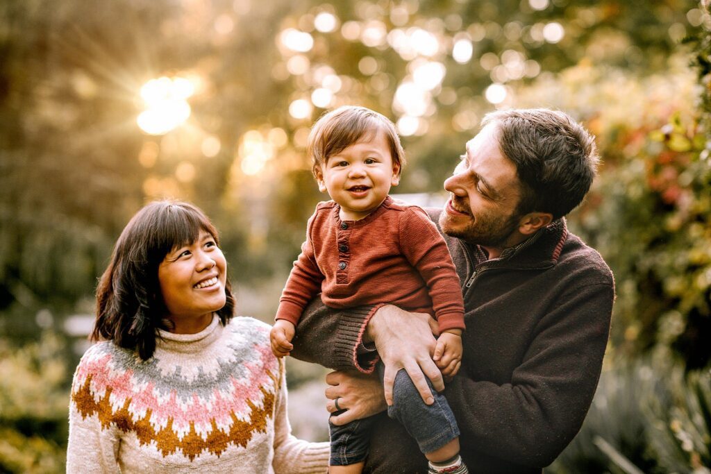 A smiling couple stands outdoors with a young child between them. The man holds the child, who is smiling—a sign of a successful photoshoot. The woman looks at them fondly as sunlight filters through trees in the background, capturing a beautiful moment from their kids photoshoot.