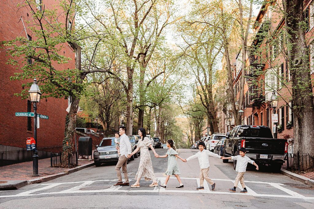 A family of five, having spent time preparing kids for a photoshoot, holds hands as they cross a tree lined city street at a crosswalk, surrounded by parked cars and brick buildings on a sunny day.