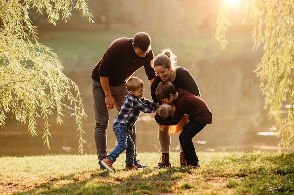A family of four, two adults and two children, play together outdoors on a grassy area by a pond, with sunlight filtering through trees in the background. Their laughter fills the air as they enjoy these precious moments—perfect for a kids photoshoot and capturing natural smiles. This relaxed setting is one of the successful photoshoot tips that helps in preparing kids for photoshoot sessions, ensuring everyone feels comfortable and.